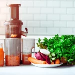 Estrattore di succo a freddo in cucina con un bicchiere di succo di carota e un piatto di verdure crude assortite (carote, barbabietole, rapa bianca, cetriolo, prezzemolo ed erbe aromatiche) sul piano di lavoro.
