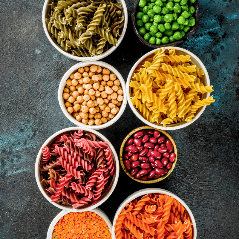 Un assortimento di paste e legumi colorati in ciotole individuali. Sono visibili pasta di piselli, ceci, pasta di grano saraceno, fagioli rossi, lenticchie rosse e pasta di lenticchie, allineate su un piano scuro.